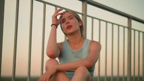 tired young lady sitting outdoors near iron railing at dusk wipes her hand and forehead, takes deep breath showing exhaustion from long day, wearing black wrist bangle and light blue tank top