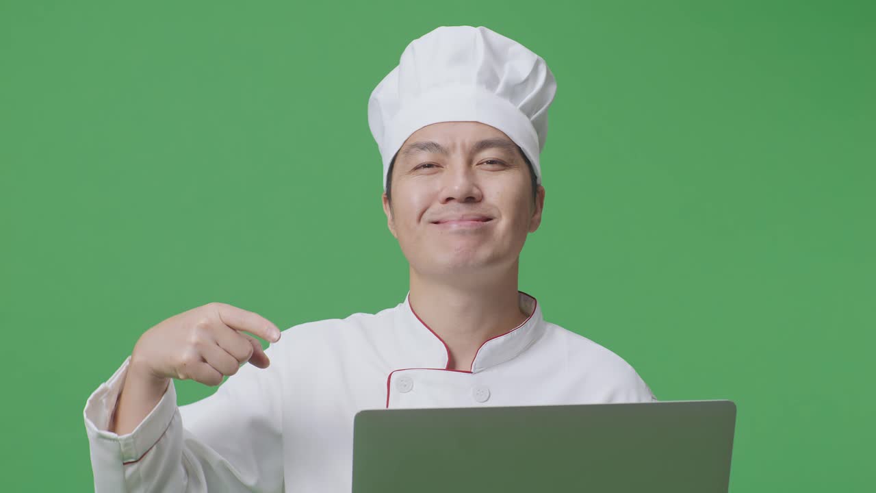 녹색 화면 배경 스튜디오에 서 있는 동안 손에 노트북을 가리키며 미소 짓는 아시아 남자 셰프의 클로즈업