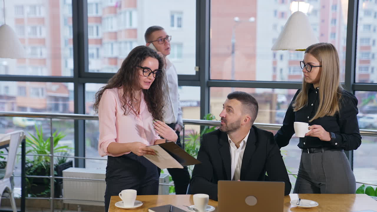 Bearded man sitting at table listens to female colleagues standing at both sides. Man talking on the phone at backdrop.