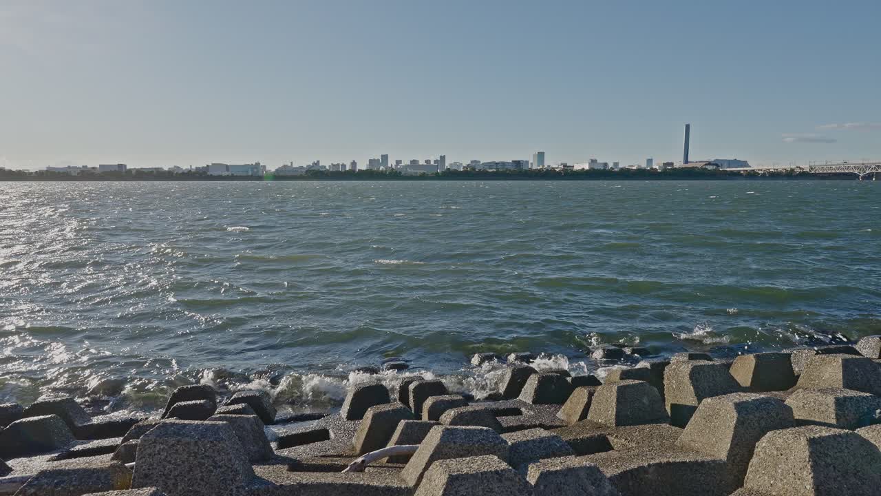 Daytime shot from behind large concrete tetrapods toward a choppy bay with a distant city skyline and industrial stack