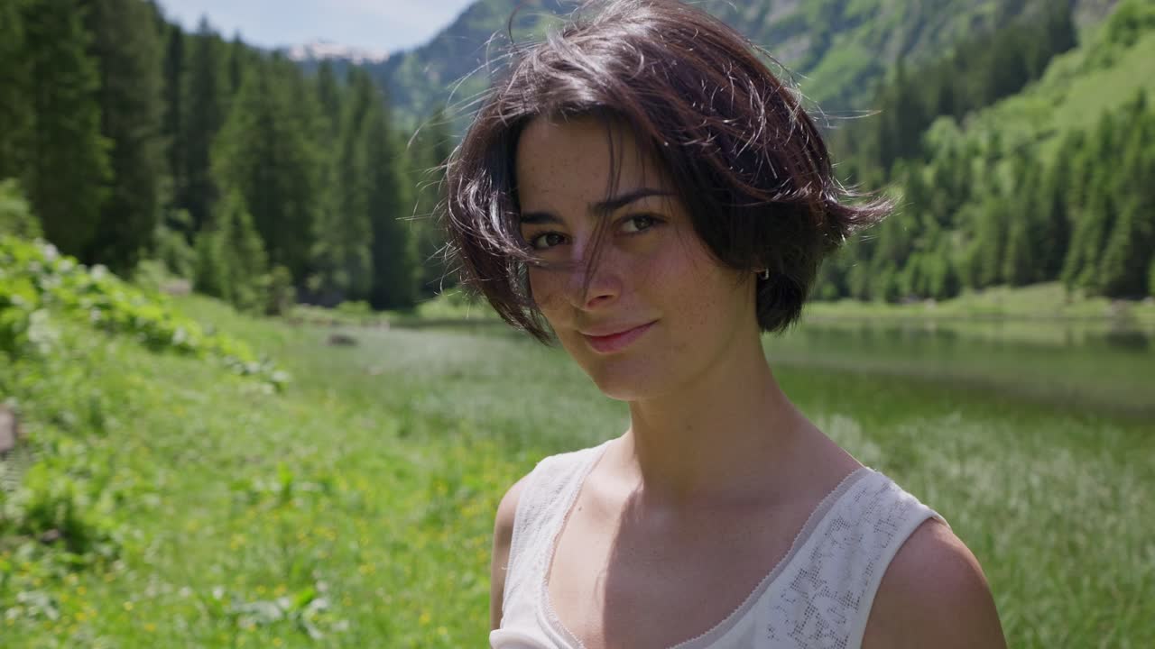 A young woman runs her hand through her hair by a lake in a lush mountain setting