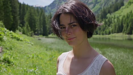 a young woman runs her hand through her hair by a lake in a lush mountain setting