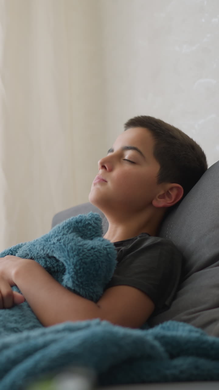 Boy suffering from flu lies on couch with thick blanket covering him, coughing and covering mouth in softly lit room near window with sheer curtain, expressing fatigue