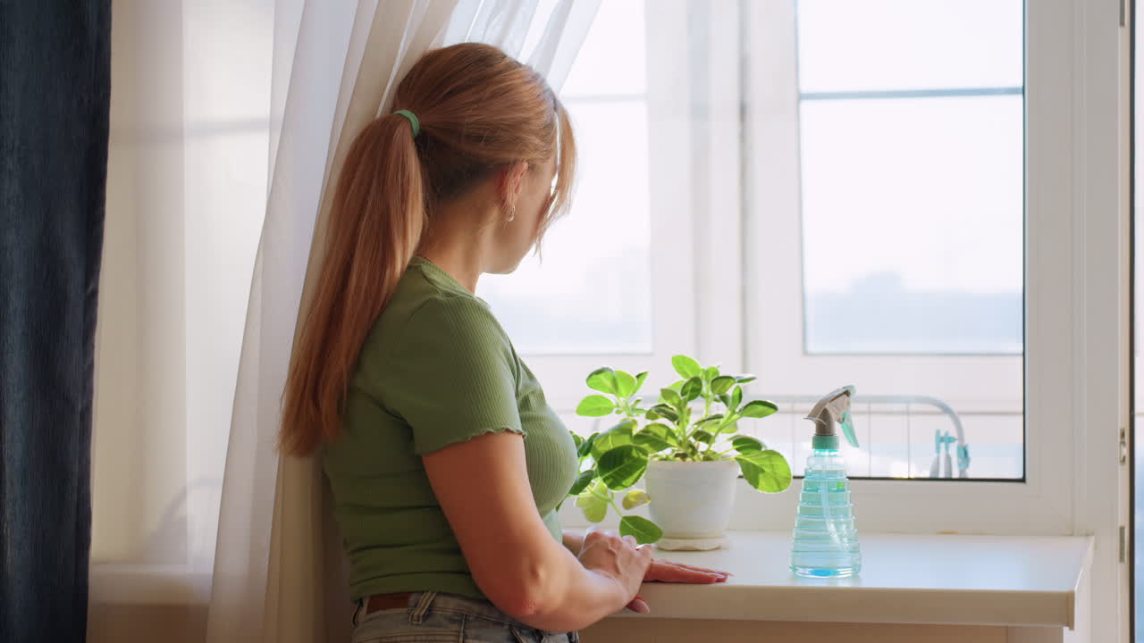 Young woman with ponytail in green shirt standing near window, gazing outside with thoughtful expression, potted plant and blue spray bottle on windowsill