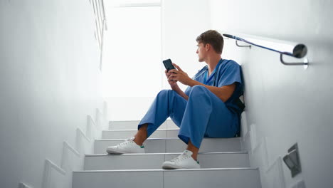 Krankenpfleger-Oder-Arzt-In-Kitteln,-Der-Auf-Dem-Mobiltelefon-Surft-Und-Auf-Der-Treppe-Im-Krankenhaus-Sitzt