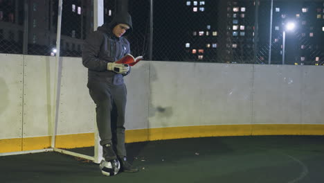 athlete in hoodie leaning against metallic goalpost reading book outdoors at night with a soccer ball underfoot, illuminated by urban city lights