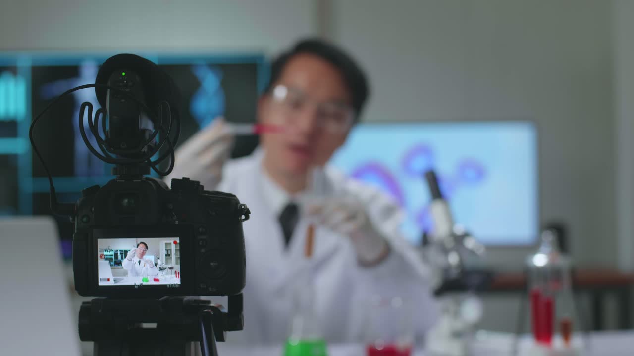 Camera Display Of Young Asian Male Research Scientist Holding Glass Test-Tube And Explain