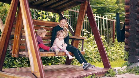 travelling young pretty mother with children swinging on a wooden swing near the summer house on vacation 4