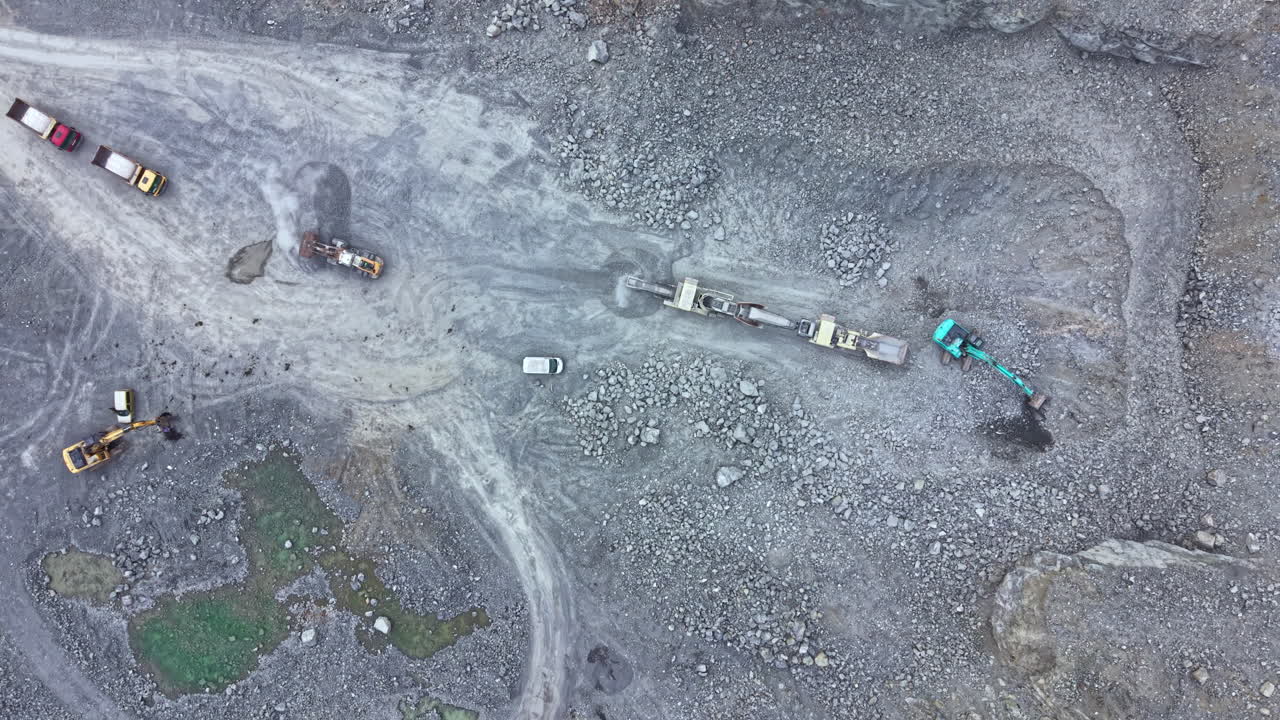 Construction work takes place at a gravel site with heavy machinery moving rocks near a dirt path in an industrial area during daylight hours