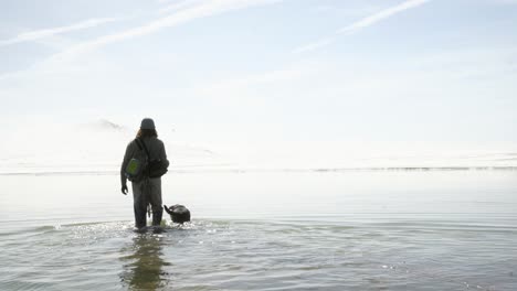 High-key-background:-Fly-fisherman-and-dog-walk-into-pale-blue-lake