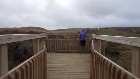 tracking follow shot of happy girl walking on the wooden bridge near the edge of a cliff with hills in the background on a cloudy day in scotland, isle of skye