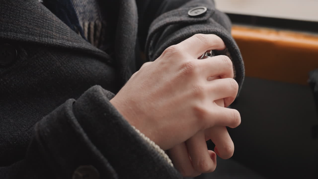 Creator hands clasped on bus seat, closeup of fingers fidgeting inside wool coat sleeve, button detail, window light, quiet commuter mood, planning next project with nervous energy
