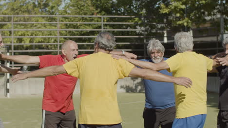 felices jugadores de fútbol senior celebrando la victoria, saltando en círculo y abrazándose en el campo