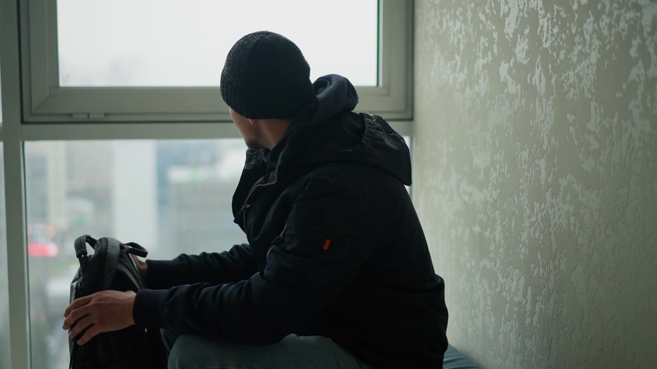 Close up of man in dark clothing sitting by window holding black backpack and looking out. Indoor setting with textured wall visible soft lighting from outside through frosted glass panels