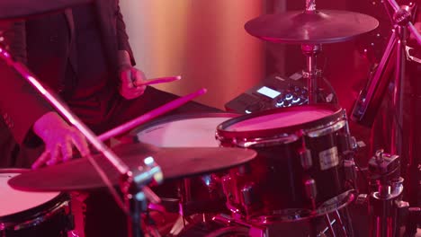 drummer playing a drum kit under red lighting during a live performance.