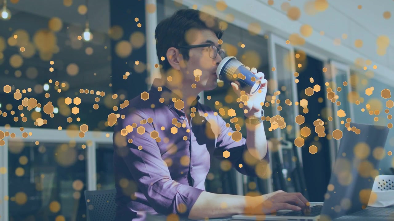 Uomo che solleva una tazza di caffè, sorseggiando mentre digita sul portatile per concentrarsi sul lavoro, con bokeh dorato in movimento