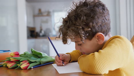 niño joven en casa con un ramo de flores escribiendo en la tarjeta del día de la madre