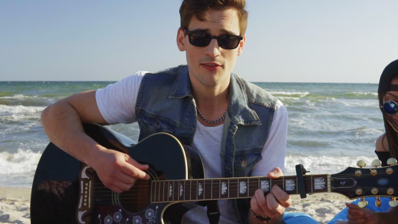 joven tocando la guitarra entre un grupo de amigos sentados en sillas fáciles en la playa y cantando en una noche de verano. disparo en cámara lenta
