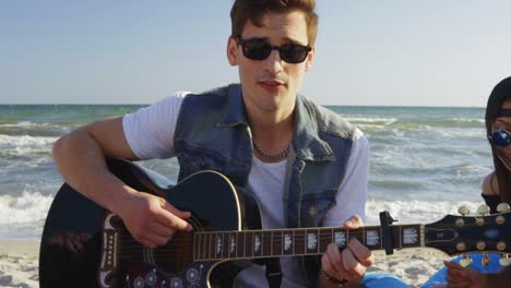 young man playing guitar among group of friends sitting on easychairs on the beach and singing on a summer evening. slowmotion shot