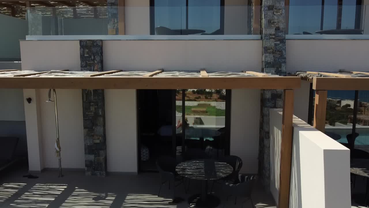 수영복을 입은 갈색 여성은 그리스 크레타의 카이오 리조트의 수영장에서 물을 즐기고 테일을 마시고 드론이 날아와 리조트를 볼 수 있습니다.