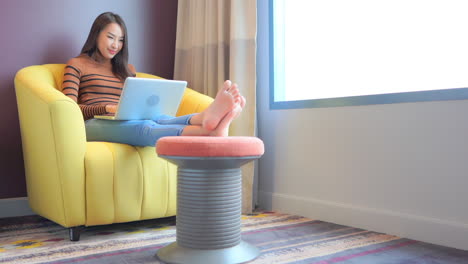 asian woman typing on laptop keyboard while sitting in yellow armchair having her legs resting on the puff at apartment room