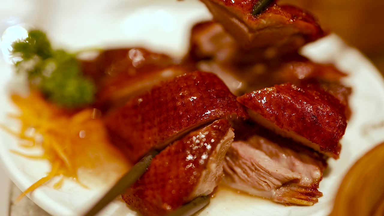 Close-up of roast duck being served with chopsticks at a Chinese restaurant. Warm lighting enhances the rich, glossy texture