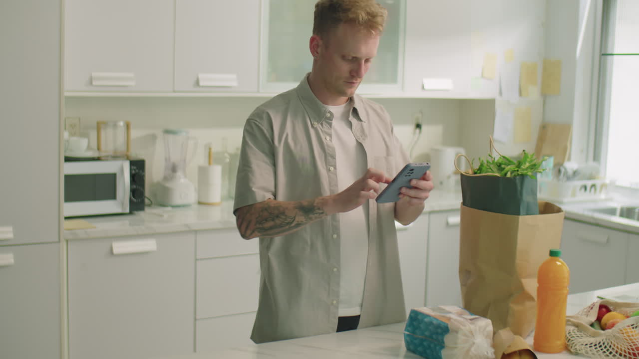 Man Standing by Groceries on Kitchen Table and Using Phone
