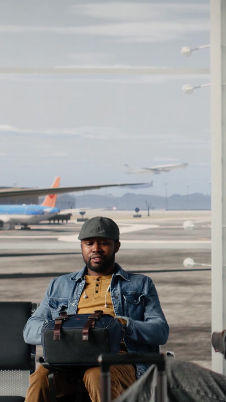 Uomo in attesa nella sala partenze dell'aeroporto con il tabellone delle partenze e aerei sullo sfondo.