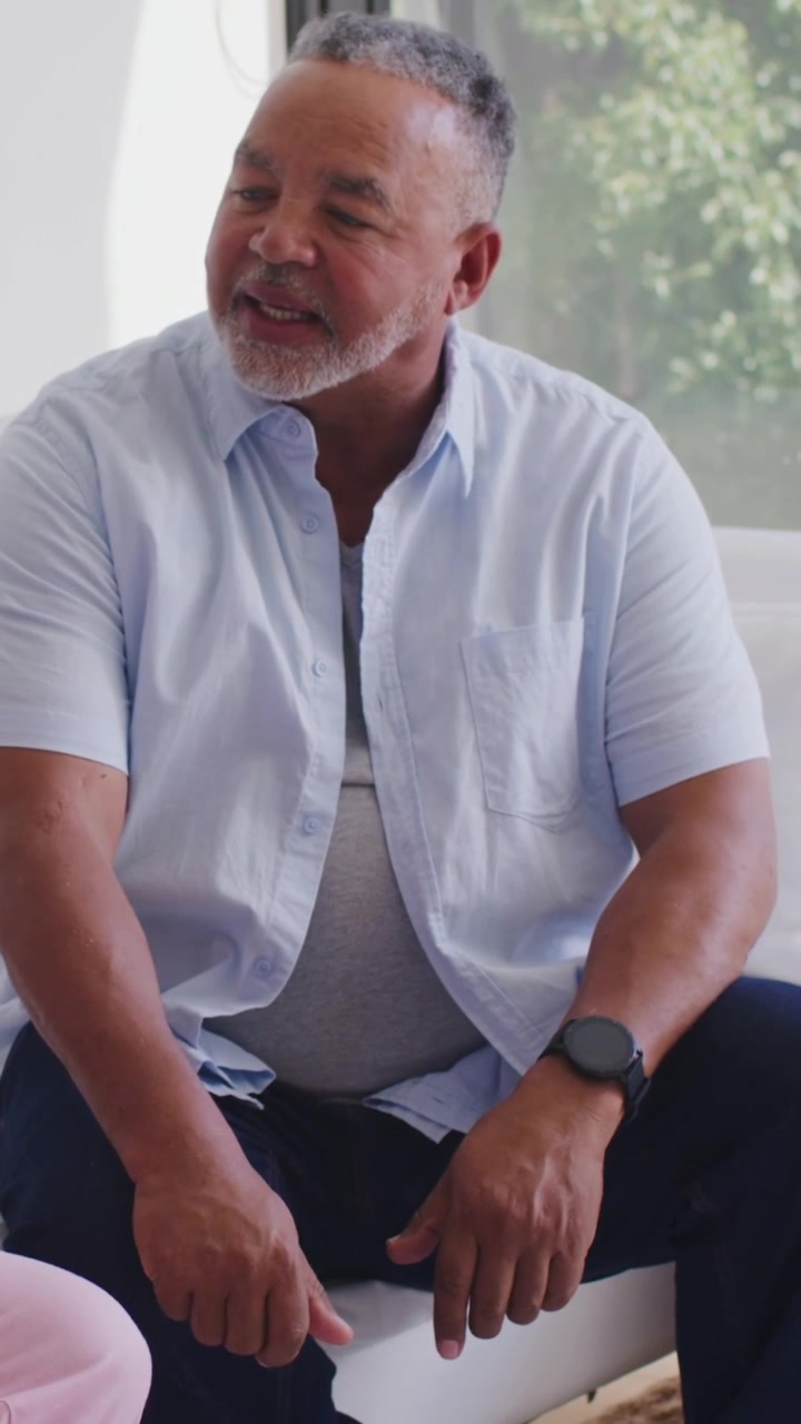 Video verticale: Compagno che parla, anziano seduto con camicia blu che ascolta, battendo sull'orologio vicino alla finestra