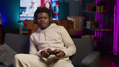 young man sitting on sofa at home relaxing by playing videogames on gaming console, unwinding in neon lit apartment by competing in online multiplayer e sports tournament. camera a.