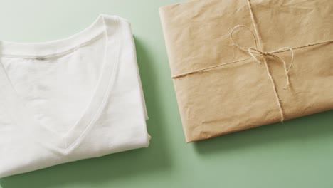 Video-of-close-up-of-white-folded-t-shirt-with-brown-paper-parcel-on-green-background