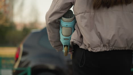 close-up shot of a girl in a peach jacket and black pants holding a turquoise skateboard, with her long brown hair visible, standing still on a park pathway as a car approaches in the background