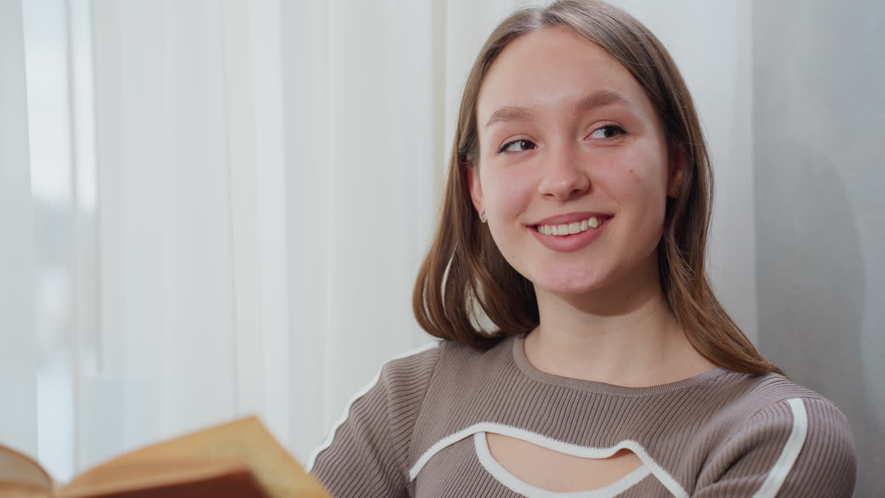 갈색 표지 책의 페이지를 뒤집는 여자가 따뜻한 미소로 쳐다보고, 투명한 유리창 에 앉아, 부드러운 자연 빛이 평화로운 분위기에서 편안한 실내 공간을 조명합니다.