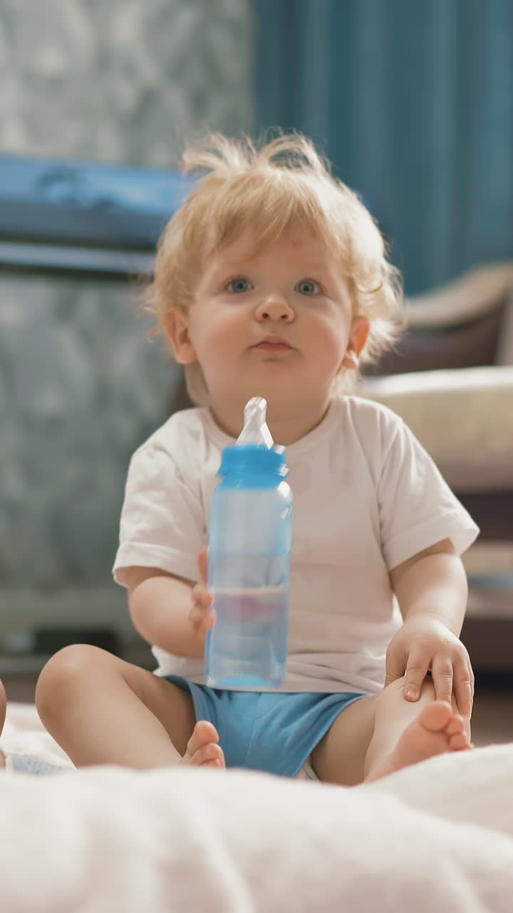 camera tracking low angle: children sit on floor, girl plays on smartphone, boy drinks water from bottle through pacifier, smiling, with blond hair, under sunlight