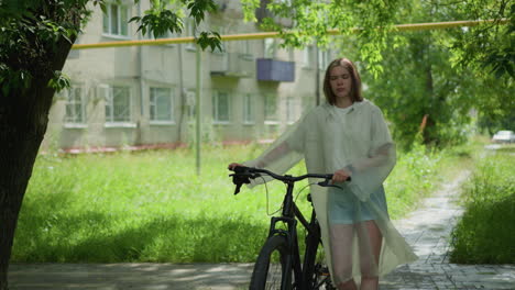 thoughtful woman in raincoat with blonde hair walking her bicycle on paved path, holding black object in right hand, background includes lush greenery, trees, building, and yellow pole