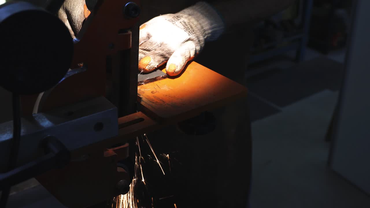 Man using protective glove sands a knife using a sanding machine creating red sparks with trails