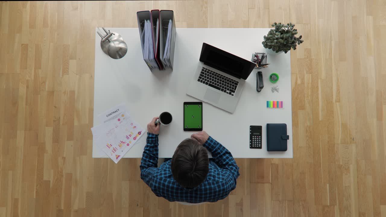 Top view shot of an office worker sitting on desk using tablet with greenscreen chroma key drinking coffe with floating sun