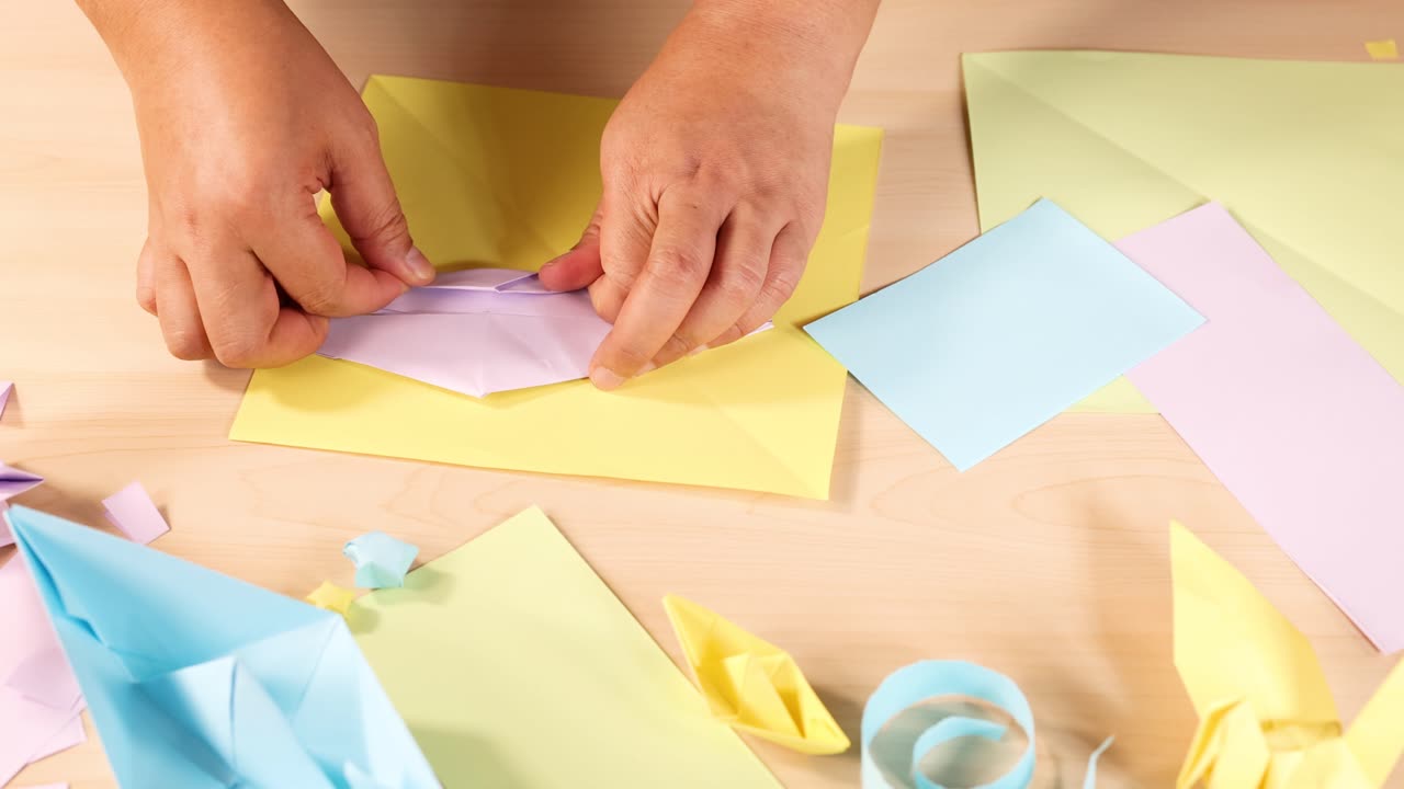 Person folds pastel origami paper, surrounded by colorful crafts, under bright, even lighting overhead