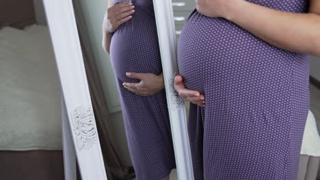 future mother with advanced pregnancy comes up to big mirror. woman in a long nightgown in front of the big mirror caressing the tummy with two hands. close up.