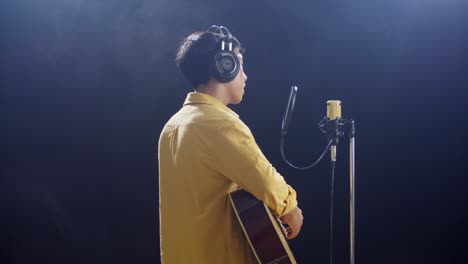 side view of young asian boy with headphone playing a guitar and singing into a condenser microphone on the white smoke black background