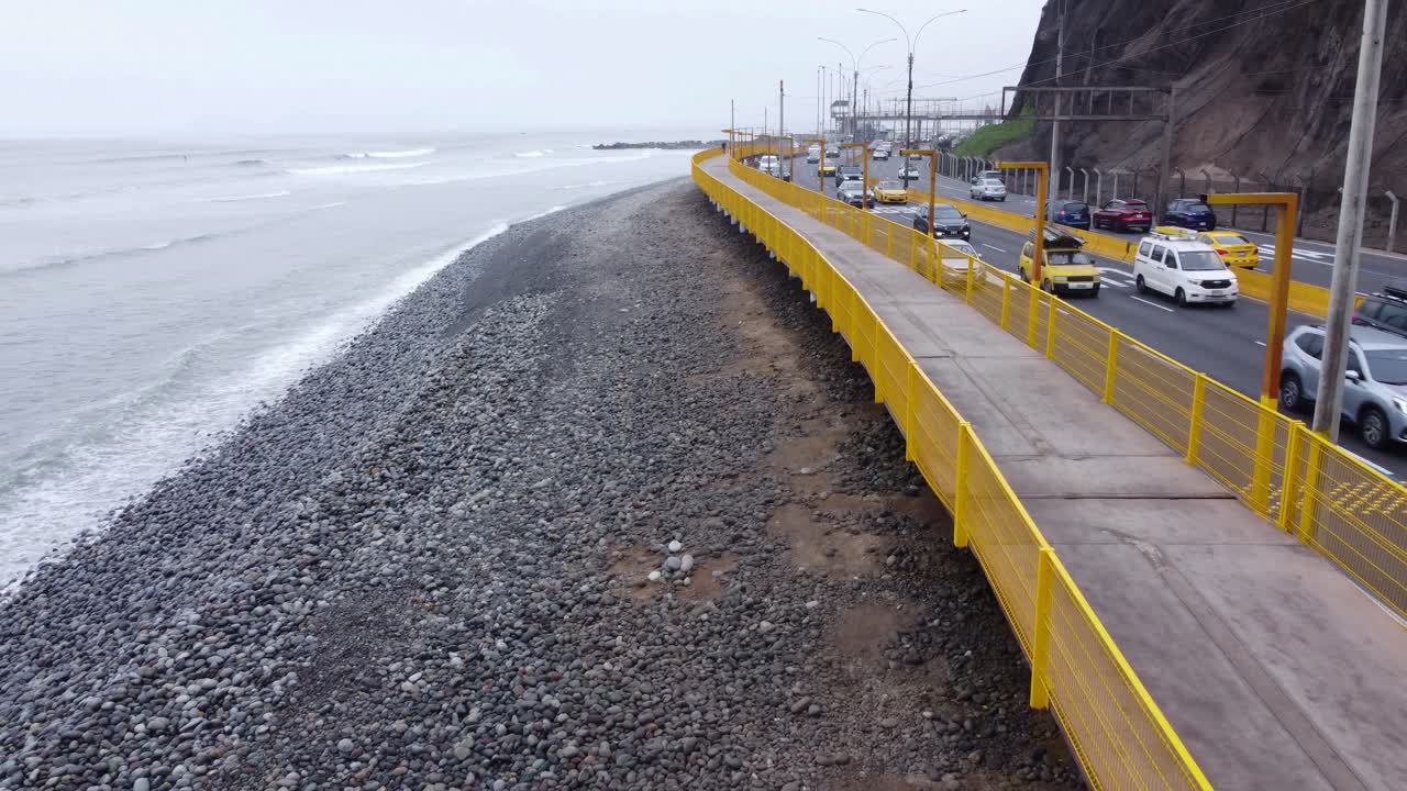 Coastal freeway with the rocky beach on one side and tall cliffs on the other. Cars drive by as the drone flies forward next to walking path. Located in the Miraflores district of Lima, Peru