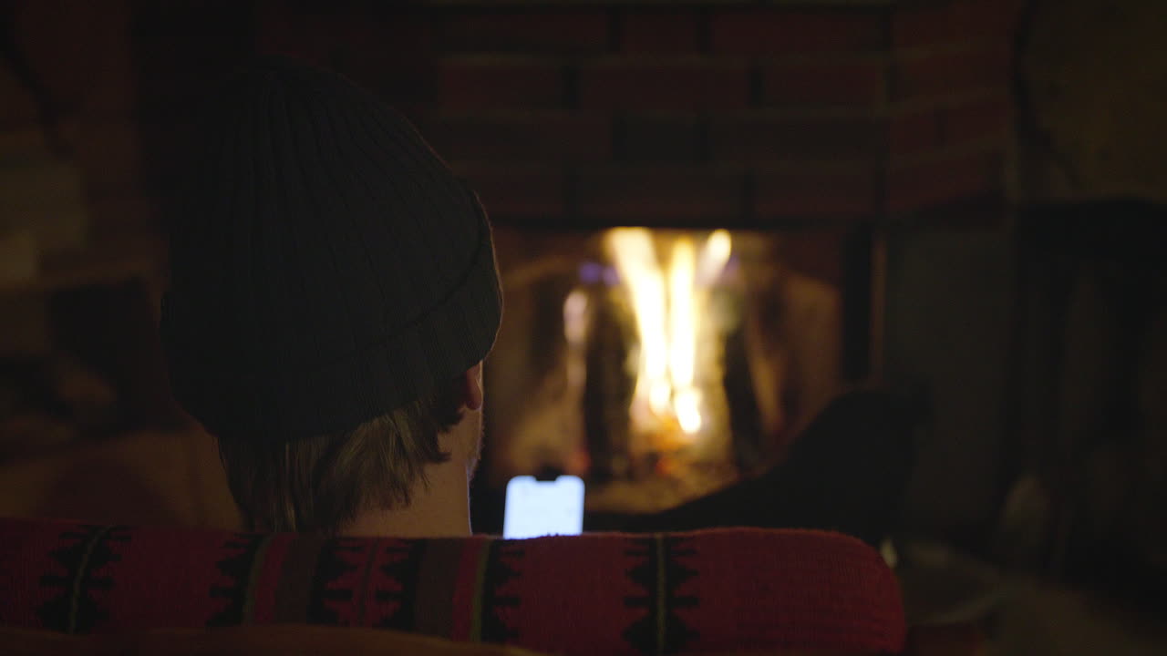 A person in a knit beanie and warm sweater sits near a glowing fireplace, holding a smartphone in a dimly lit cozy indoor setting. Norwegian cabin setting.