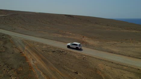 Vista-Aérea-Lateralmente-Siguiendo-Un-Coche-Blanco-En-Una-Carretera-Volcánica-Y-Desértica-En-La-Isla-De-Fuerteventura-En-Un-Día-Soleado