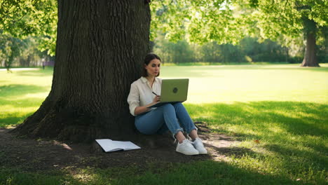 young woman working on laptop under a tree in a sunny park