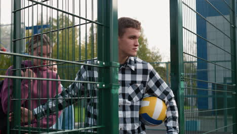 coach in plaid shirt with a volleyball under his arm opens a green gate, followed by students with a basketball court and trees visible in the background