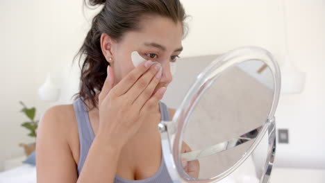 Focused-biracial-teenage-girl-putting-on-under-eye-masks-looking-in-handheld-mirror,-slow-motion