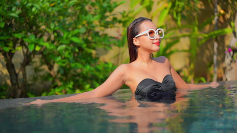 Lächelnde-Glückliche-Asiatische-Dame-In-Einem-Swimmingpool,-Die-Einen-Schwarzen-Bikini-Und-Eine-Weiße-Sonnenbrille-Trägt