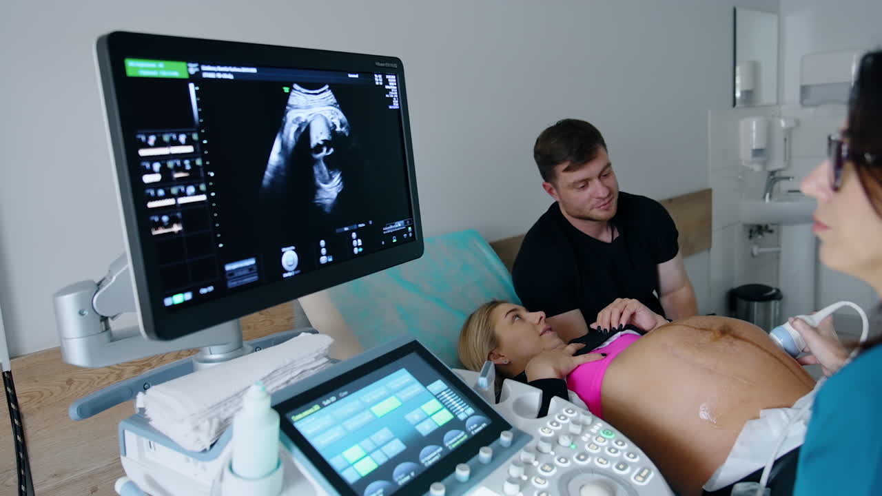 Female doctor looks at the monitor of ultrasound machine and comments the image. Caucasian couple expecting a baby at backdrop.