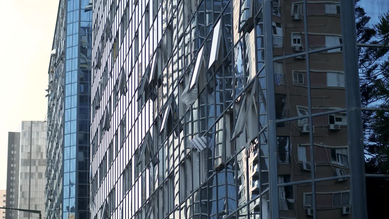 Glass-fronted tower in Tel Aviv reflects a nearby damaged building with shattered windows after a recent strike.