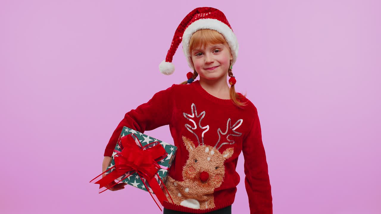 산타 스웨터를 입은 즐거운 소녀가 크리스마스 선물 상자 두 개를 선물하고 카메라로 손을 펴고 있습니다.